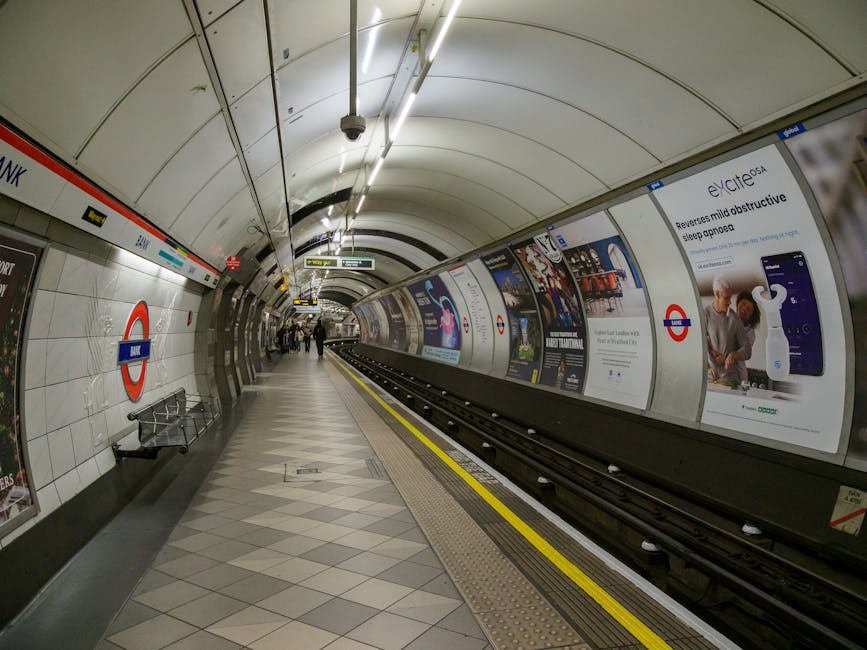 A view of Elm Park underground station platform with a curved, tiled white and beige ceiling, illuminated by bright fluorescent lights. The platform features a row of advertising boards along the wall