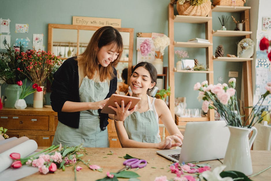 Two women inside a flower shop engaged in a discussion about floral arrangements, with one holding a tablet and the other smiling. The shop features a variety of fresh flowers including pink roses, ca
