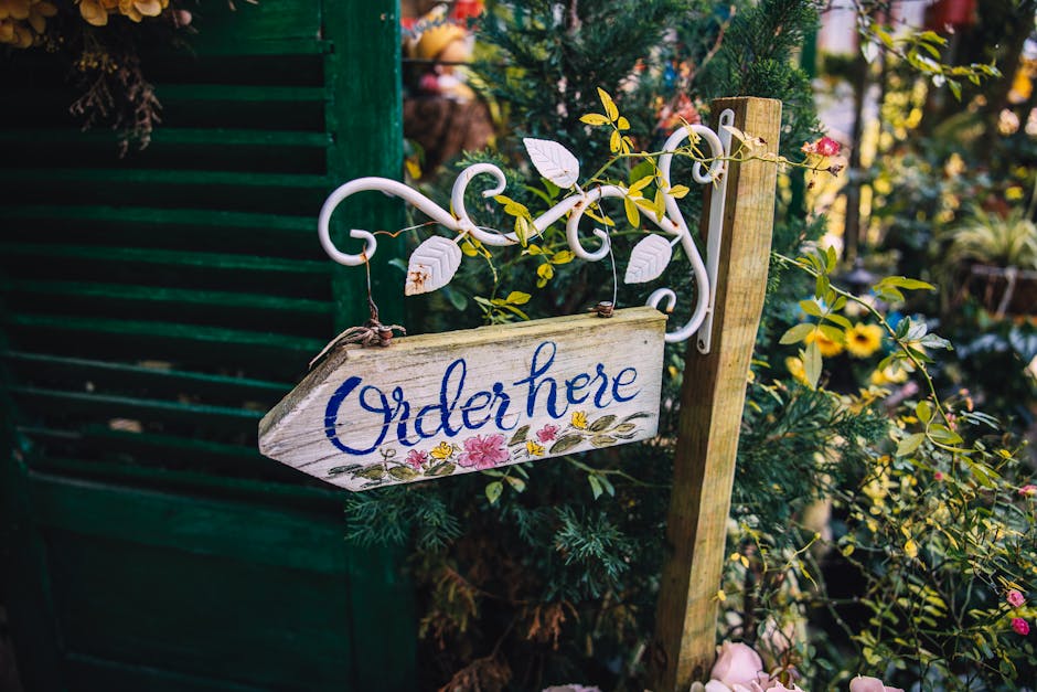 A rustic wooden sign with the words 'Order here' painted in blue cursive, hanging by metal hooks from an ornate white wrought iron bracket attached to a wooden post. The sign features a delicate flora
