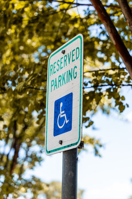 The image features a white reserved parking sign with green and blue accents, indicating accessibility with a wheelchair symbol, mounted on a metal pole. The sign is positioned outdoors against a back
