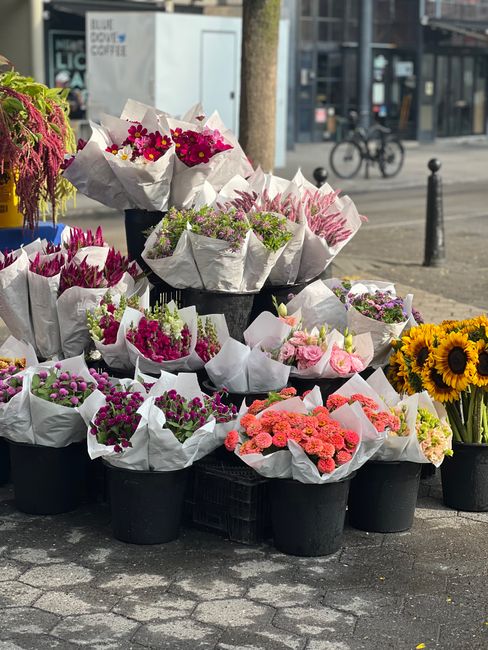 A display of fresh flower bouquets arranged in black pots and wrapped in white tissue paper sits on a paved sidewalk in an outdoor urban setting. The bouquets feature a variety of colorful flowers, in