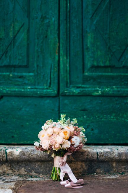 A wedding bouquet of fresh, pastel-colored flowers including cream and blush pink roses, white peonies, and delicate greenery, arranged in a rounded, lush style. The bouquet is wrapped with a soft pin