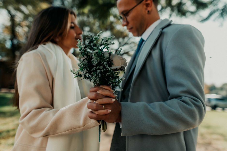 A woman in a cream-colored coat presents a bouquet of fresh flowers to a man in a light gray suit outdoors. The bouquet features a large, pale pink rose surrounded by sprigs of white and green foliage