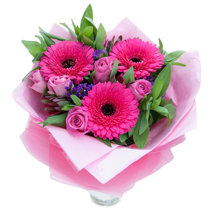 I picture this arrangement placed on a small Elm Park side table, a vivid study in pinks and texture. Three magenta-fuchsia Gerbera daisies command attention with their concentric layers of petals and deep, almost-black centres that anchor the composition. Soft lavender-pink roses nestle between them, each bloom tightly curled and velvety, lending a gentle, romantic counterbalance to the energetic gerberas. Tiny sprays of purple statice or limonium drift through the bouquet like confetti, their paper-like clusters introducing a cool violet accent and airy texture that keeps the arrangement feeling light. Lush green leaves - broad and glossy, likely salal or eucalyptus - create a sculpted backdrop, their fresh scent and crisp form giving the piece structure and a garden-sourced vitality. The selection is wrapped in an inner sheet of pale pink matte tissue and finished with a sheer, slightly crinkled fuchsia-pink cellophane that catches light and ties the whole palette together; the wrapping is folded with careful, artisan detail. The clear glass vase peeks out at the bottom, a promise of immediate display and simple elegance. Under soft daylight the petals gleam and the colours read true: energetic, thoughtful, celebratory. I imagine this bouquet finding a home after a visit to Hornchurch or as a gift delivered across Elm Park Avenue to celebrate a birthday, anniversary, or a small victory. The overall feeling is one of affectionate exuberance - thoughtfully made, seasonally fresh, and ready to bring warmth to any room.